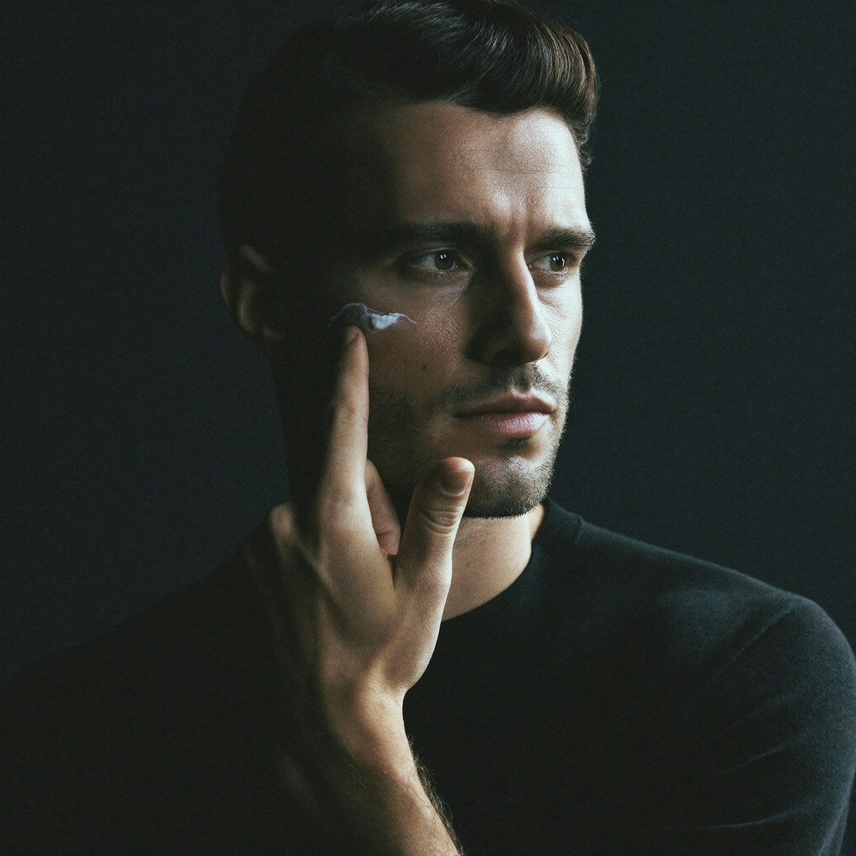 Man applying moisturiser to his cheekbone, lit against a dark background